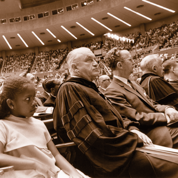 Sepia toned photo of a stadium. A young girl, an older man in a robe, and a man in a suite and eyeglasses are seated together in a row.