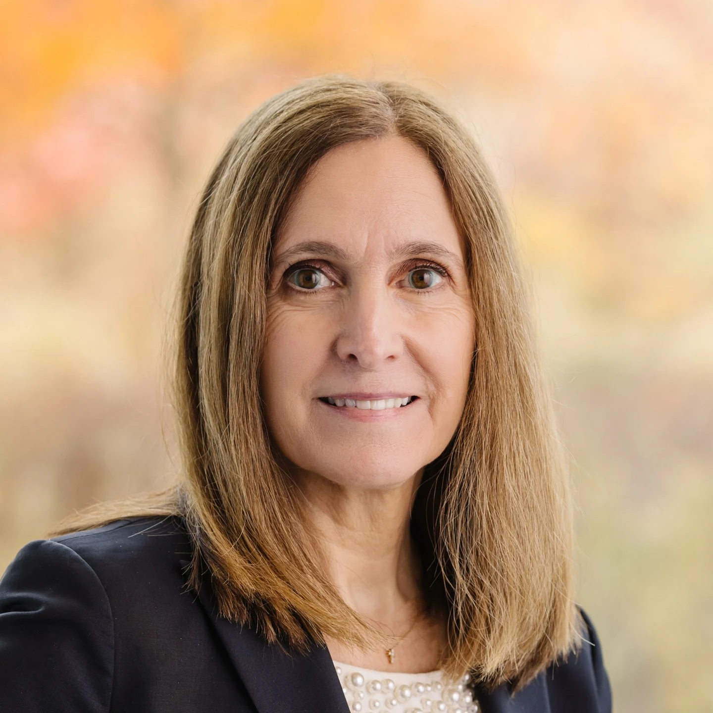 A woman with straight, light brown hair wearing a black blazer and white top smiles at the camera. The background is softly blurred with warm tones of orange and yellow, suggesting an outdoor autumn setting.