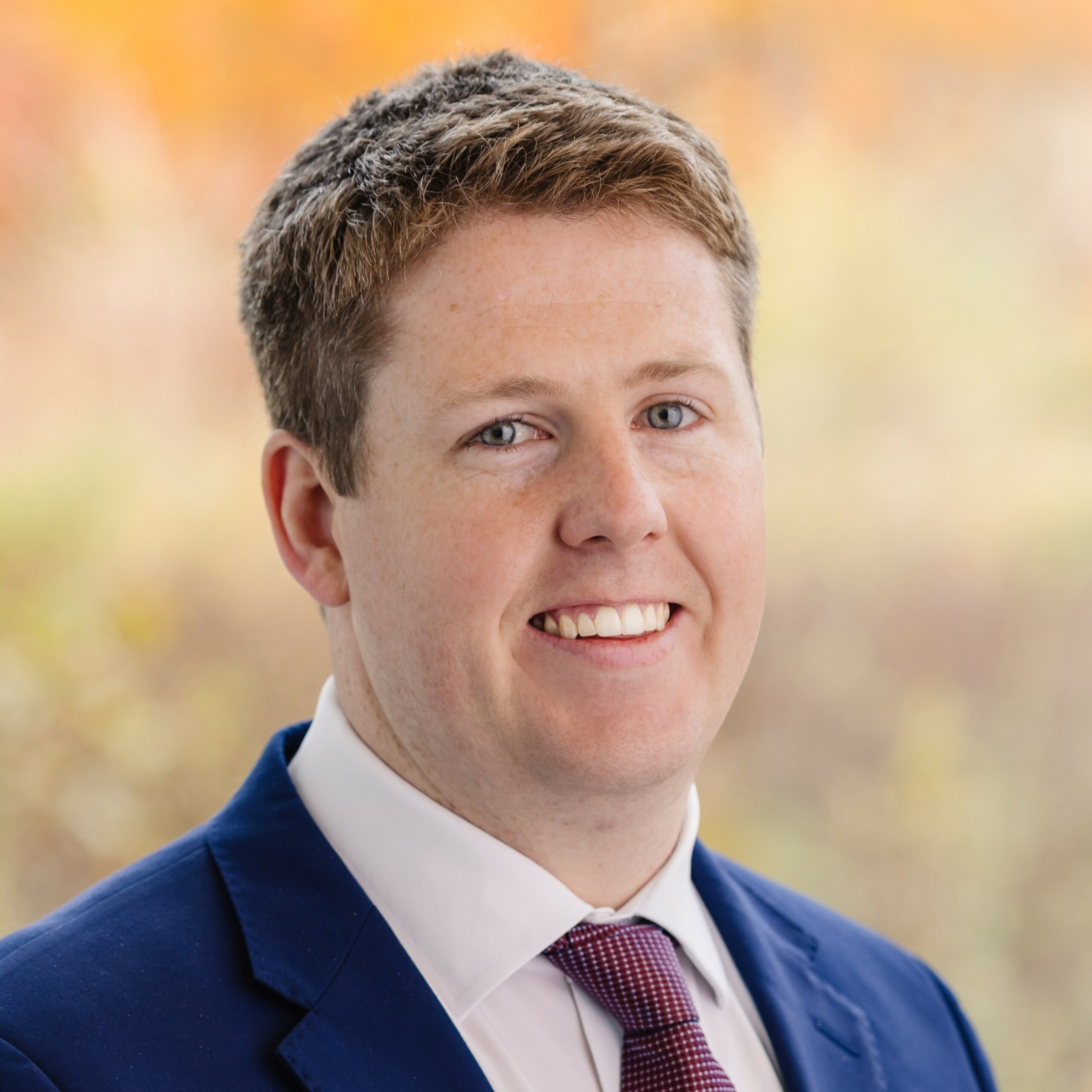 A man in a blue suit, white shirt, and red tie smiles at the camera with a blurred outdoor background of fall colors.