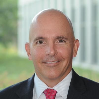 A bald man in a dark suit, white shirt, and patterned red tie smiles at the camera. The background is blurred, showing greenery and a building with large windows.