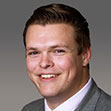 Portrait of a smiling man with short brown hair, wearing a light-colored dress shirt, tie, and gray suit jacket, set against a plain gray background.