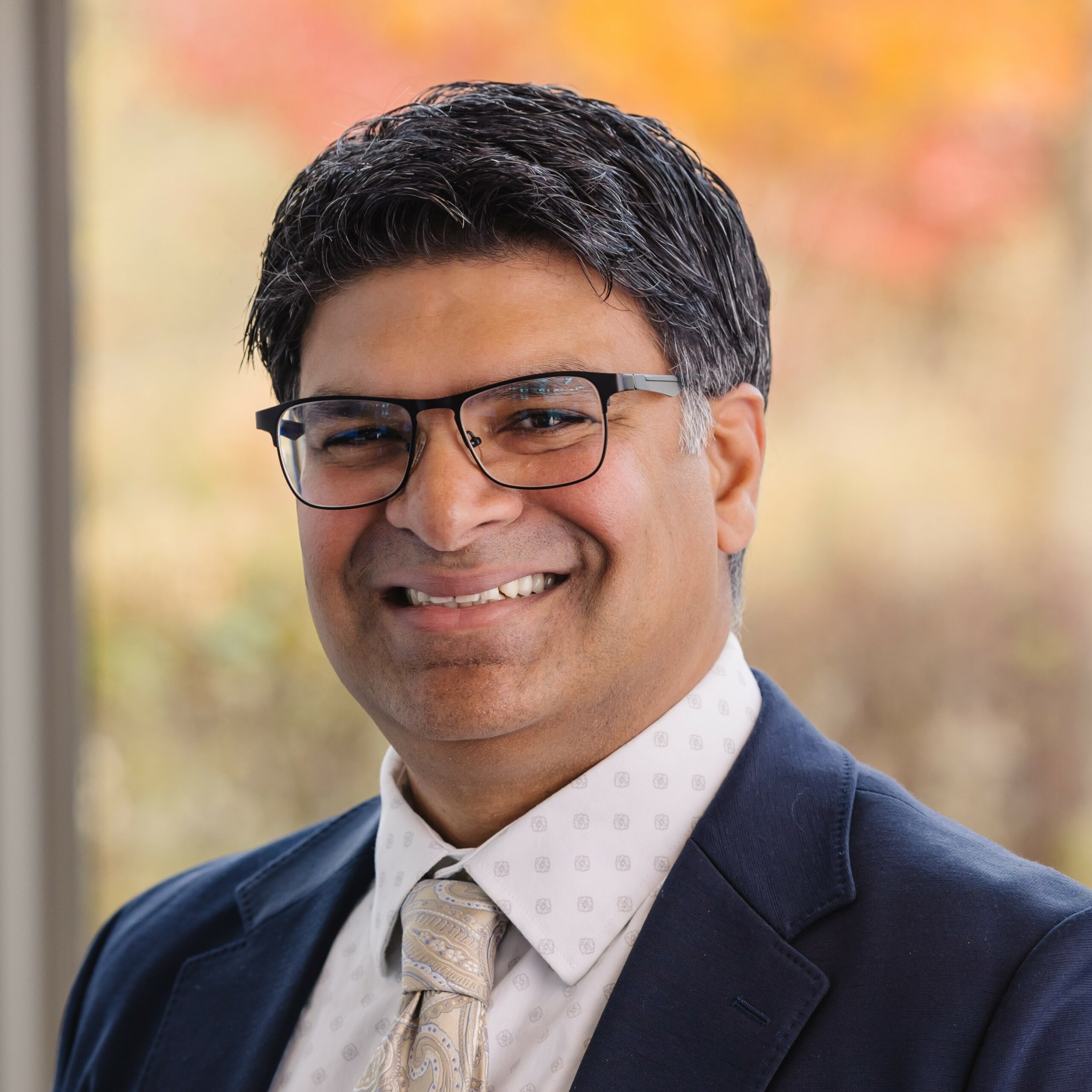 A man with short dark hair and glasses smiles at the camera. He is wearing a dark suit, a white dress shirt with a subtle pattern, and a light-colored patterned tie. The background is softly blurred with autumn colors.