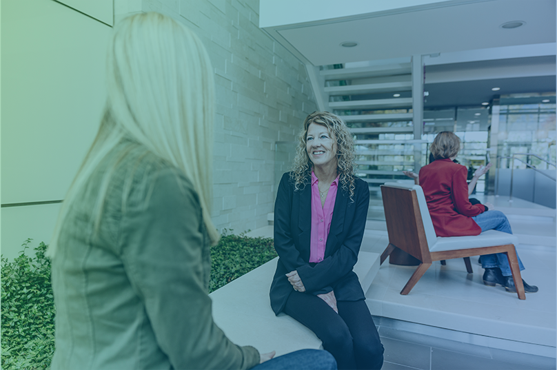 A woman is a suit sitting down smiling at a woman sitting across from here in a company lobby.