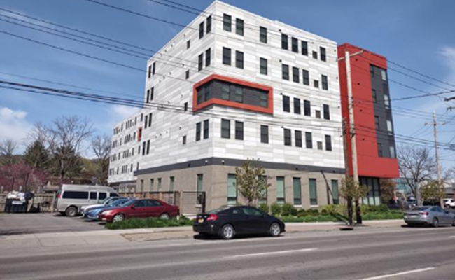 A six story building. The building has windows, and is white with grey and red accents. There are cars parked in front of the building.