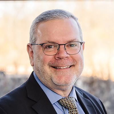 A portrait of Bryan Mjaanes. He's smiling and wearing a suit, tie and eyeglasses.