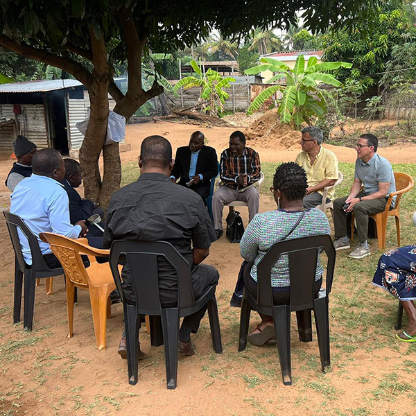 A group of men sitting on chairs arranged in a circle outside under a tree.