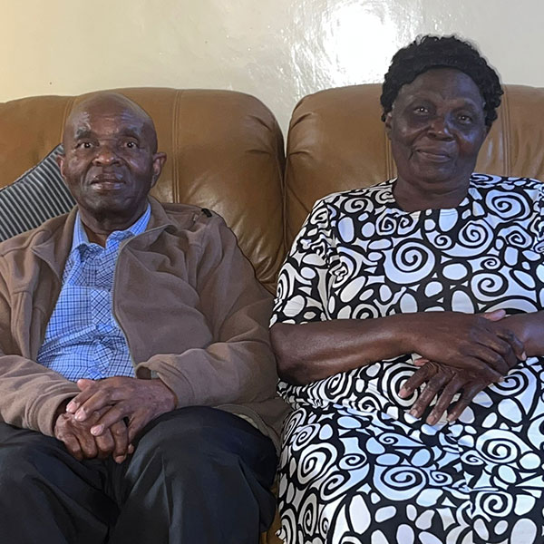 A man and a woman are seated next to each other on a tan leather sofa. The man is wearing a brown jacket, blue shirt and black pants. The woman is wearing a navy blue and white print dress.