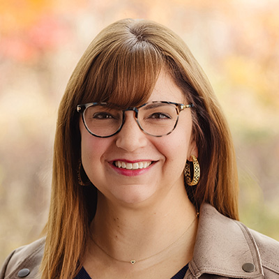 A portrait of Christina Tincher. She is wearing eyeglasses and is smiling, She has long light brown hair, a tan leather jacket, and a black blouse.