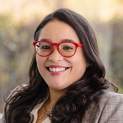 Portrait of a young woman smiling. She has long brunette hair, a tan blazer and is wearing red eyeglasses.