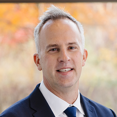 A man in a suit and tie smiles at the camera. He has short gray hair and is standing in front of a blurred background with autumn colors.