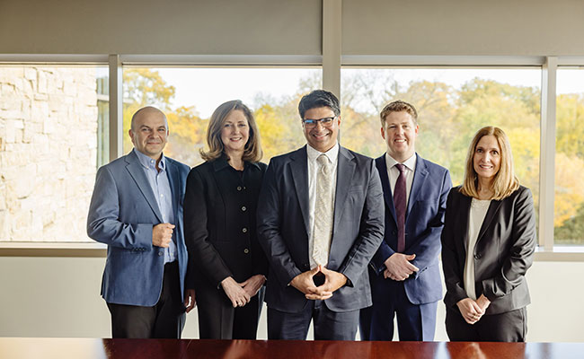 Five members of the investment team are standing in front of windows. They are dressed in business attire and are smiling.