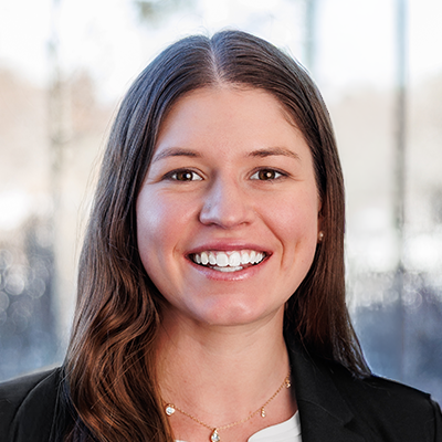 Portrait of Gabby Freeman. She's smiling, has long brunette hair, and is wearing a white blouse and black blazer.
