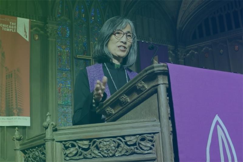A woman wearing eyeglasses and a black shirt and purple stole is standing behind a podium inside a church.