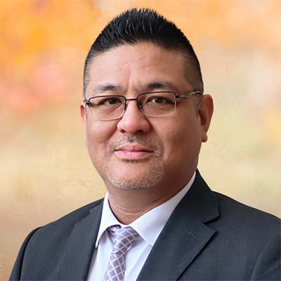 Portrait of Peter Hang smiling. He's wearing eyeglasses, a black blazer, white collared shirt and lavender tie.