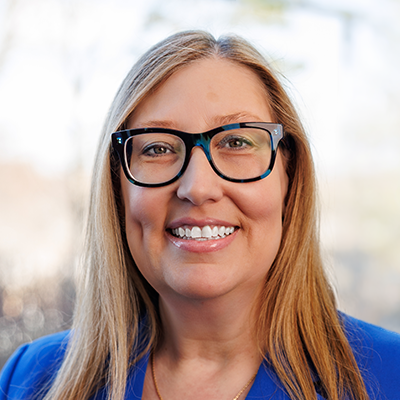 Portrait of Helen Exarhakos. She's smiling, wearing black rimmed eye glasses, and a blue dress.