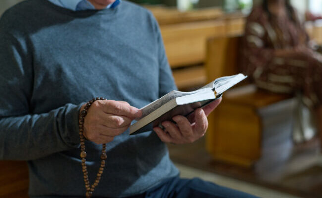 Hands of aged man holding open Bible and wooden rosary beads during sermon
