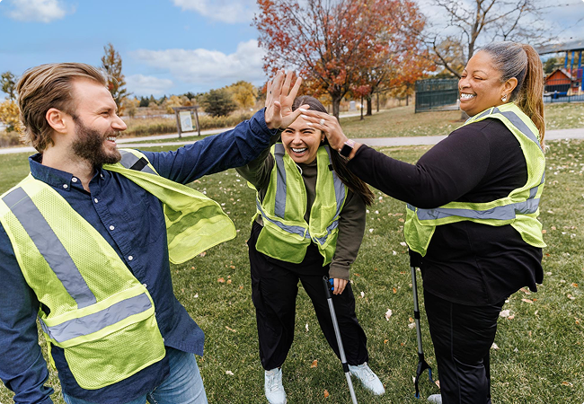 Three people in yellow safety vests smile and high-five each other while standing on grass in a park, with autumn trees in the background. They appear to be participating in an outdoor community activity.
