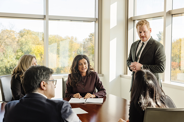 A man in a suit in standing in front of a table presenting to three team members that are seated.