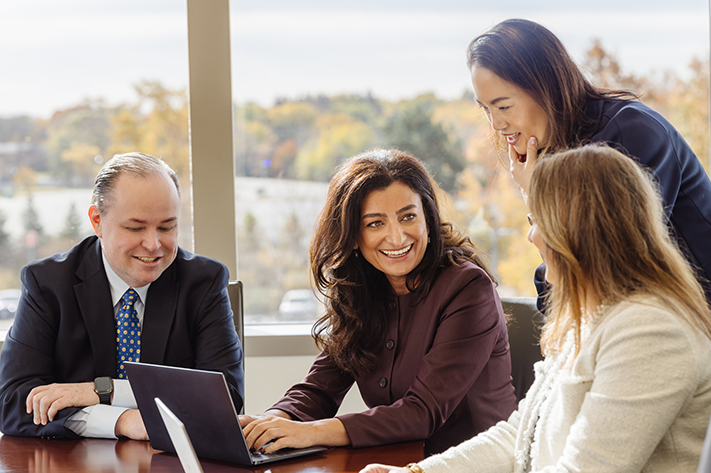 Four professionals are gathered around a table, smiling and engaged in discussion, with large windows and autumn trees in the background. A man uses a laptop while three women talk and listen attentively.