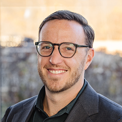 Portrait of Jake Barnett smiling. He's wearing eyeglasses, a black collared shirt and a black blazer.