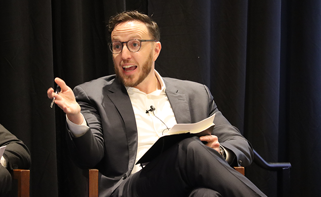 A bearded man wearing eyeglasses is seated and speaking. He's wearing a grey blazer and a white collared shirt with a microphone attached to the collar.