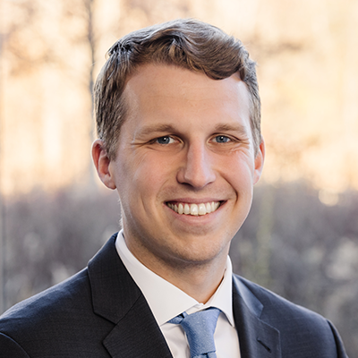 Portrait of Jon Strieter smiling. He's wearing a dark blazer, white collared shirt, and a blue tie.