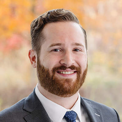 Portrait of Joshua Pullman smiling. He has a beard and is wearing a grey blazer, white collared shirt, and tie.