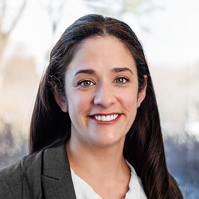 Portrait of Kate Tallo smiling. She has long brunette hair and is wearing a dark grey blazer and white blouse.