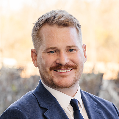 Portrait of Lucas Schoeppner smiling. He has a mustache and is wearing a blue blazer, white collared shirt and a blue tie.
