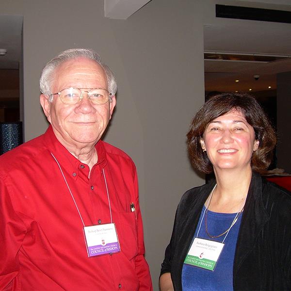 An older man wearing eyeglasses, red shirt, and lanyard with a badge is standing next to a woman with short brown hair, a black blazer, blue blouse and lanyard with a badge. Both are smiling.