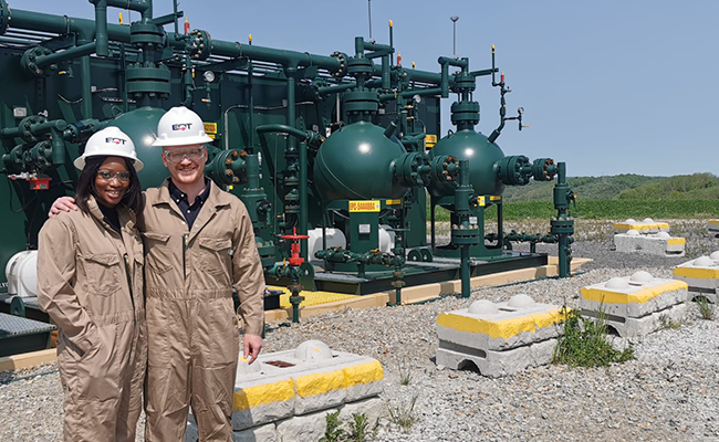 A man and a woman are standing in front of green machinery outside. Both are wearing khaki colored coveralls and white hard hats.