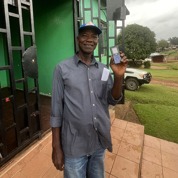 A man wearing a baseball cap, grey buttoned shirt and blue jeans. He's holding up a mobile phone.
