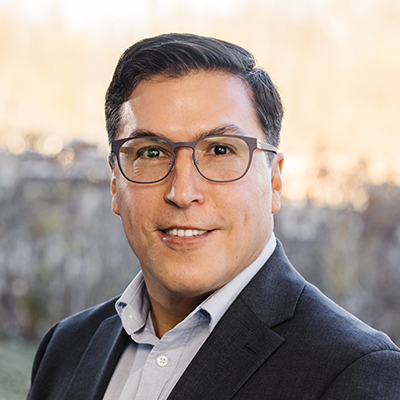 Portrait of Manuel Vargas smiling. He has short dark hair and eyeglasses. He's wearing a light blue collared shirt and a black blazer.