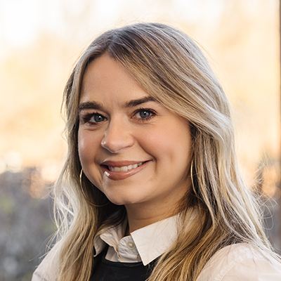 Portrait of Meagan Tenety smiling. She has long blonde hair and is wearing a white collared blouse under a black dress.