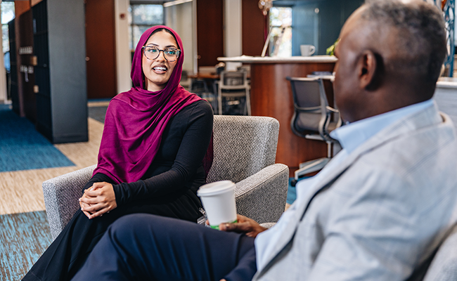 A woman wearing glasses and a purple head scarf is smiling and facing a man wearing a suit.
