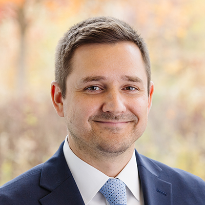 Portrait of Neil Sobczak smiling. He's wearing a navy blazer, white collared shirt and a light blue tie.