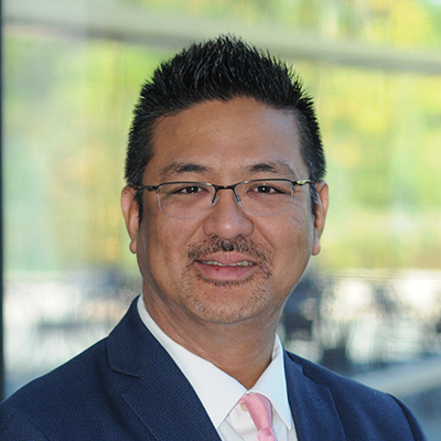 Portrait of Peter Hang smiling. He's wearing eyeglasses, a blue blazer, white collared shirt and pink tie.
