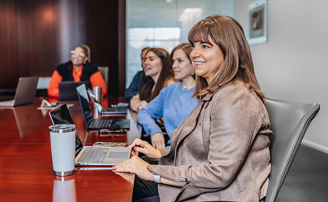 Five professionals sitting at a table during a meeting. The woman closest has her laptop open on the table and is smiling.