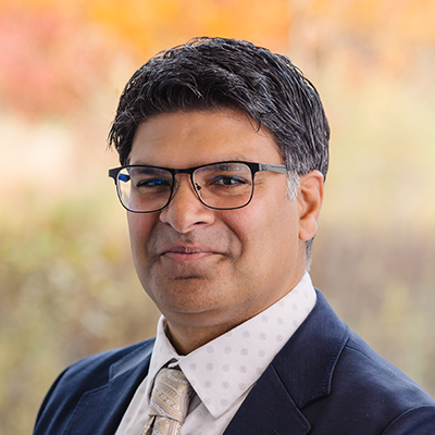 Portrait of Raj Khan smiling. He's wearing eyeglasses, a blue blazer, a light print collared shirt, and a tie.