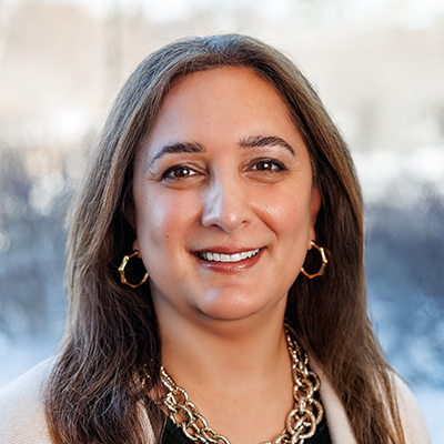 Portrait of Sheila Henretta smiling. She's wearing a gold necklace, a black blouse, and khaki blazer. She has long brown hair.