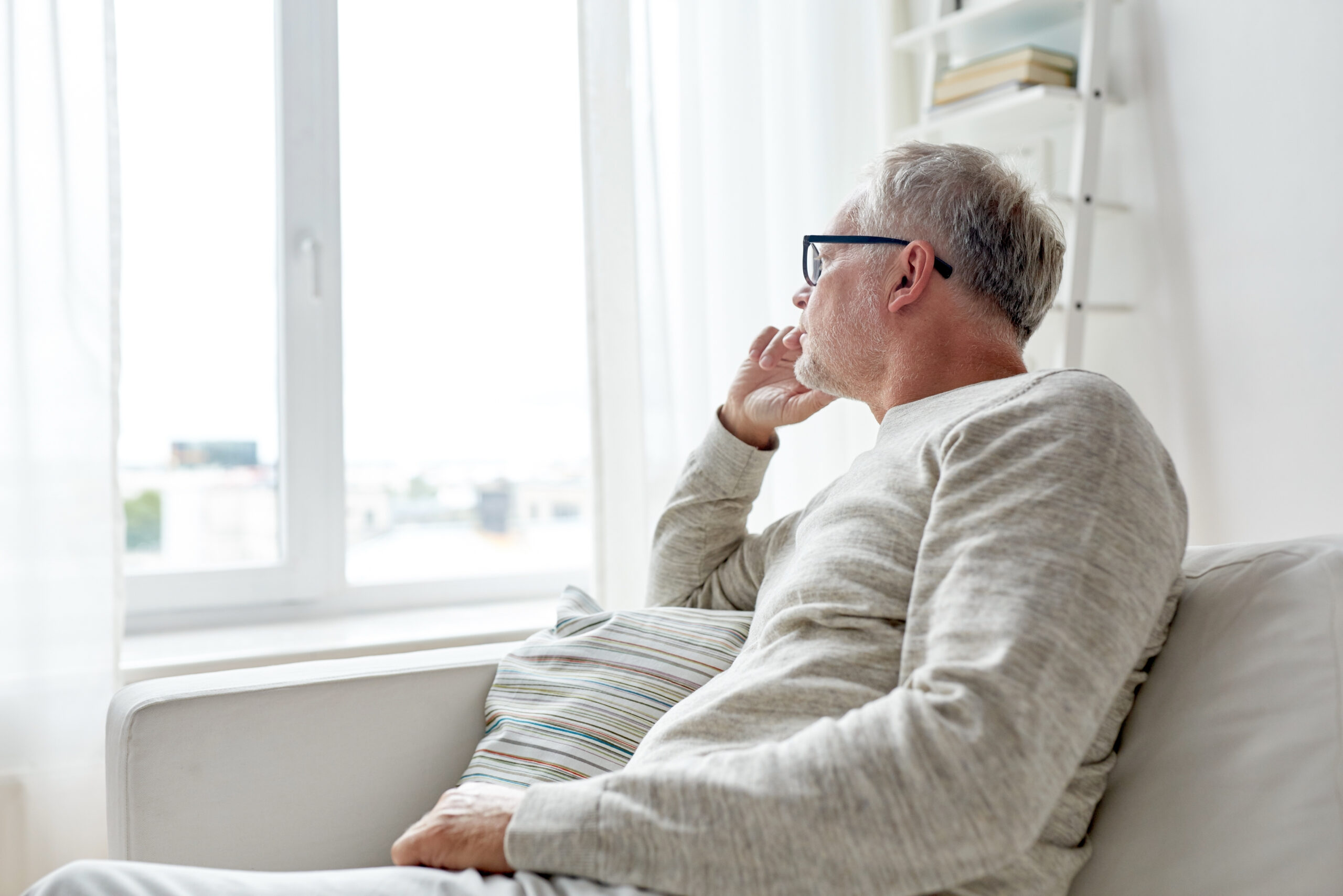 Elderly man sitting on couch looking outside window thinking