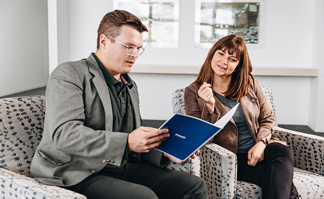 A man wearing a suit is sitting down and sharing a report with a woman wearing a brown blazer.