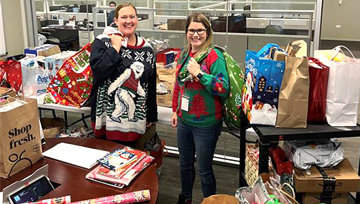Two women smiling and wearing festive sweaters. They are at a holiday gift drive. The room has tables covered with gift bags.
