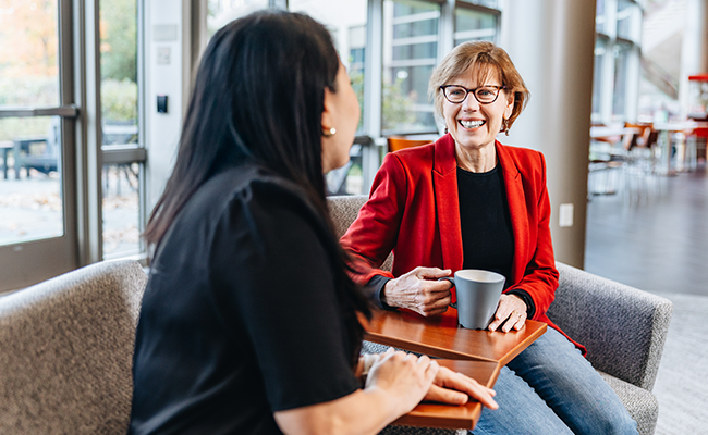 A woman with long black hair and a black blouse is seated and facing a smiling woman with short brown hair, wearing eyeglasses, a red blazer, black shirt and jeans.