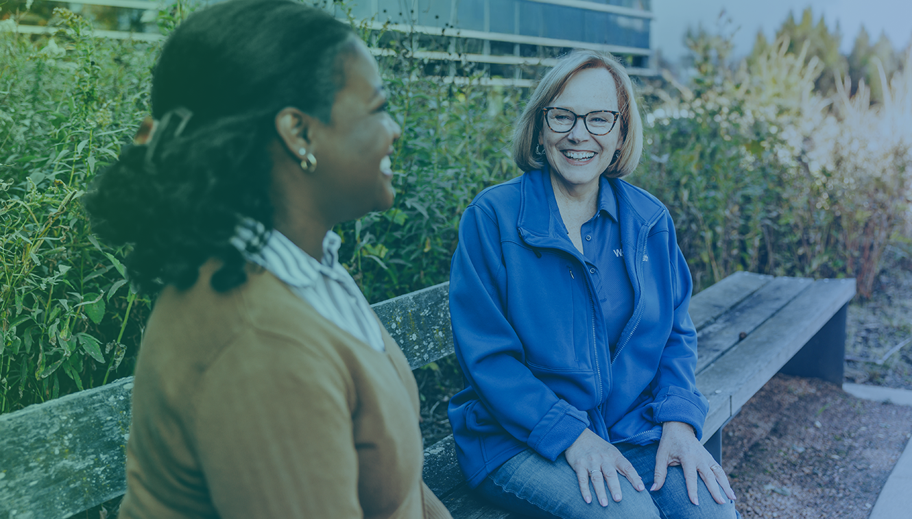 Two women sitting outside a bench smiling.