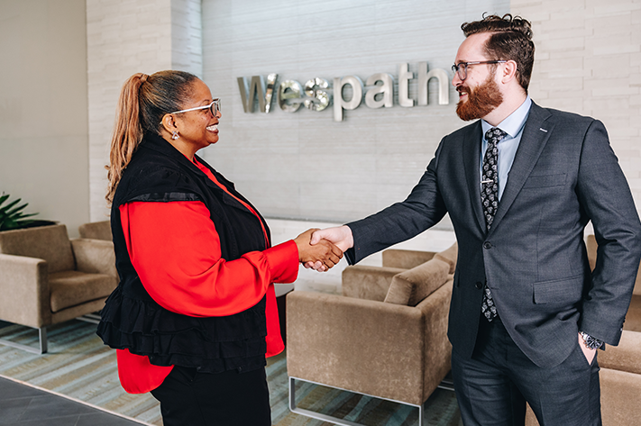 A woman wearing a red blouse and black vest is shaking hands with a man in a suit in front of the Wespath sign.
