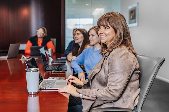 Five professionals sitting at a table during a meeting. The woman closest has her laptop open on the table and is smiling.