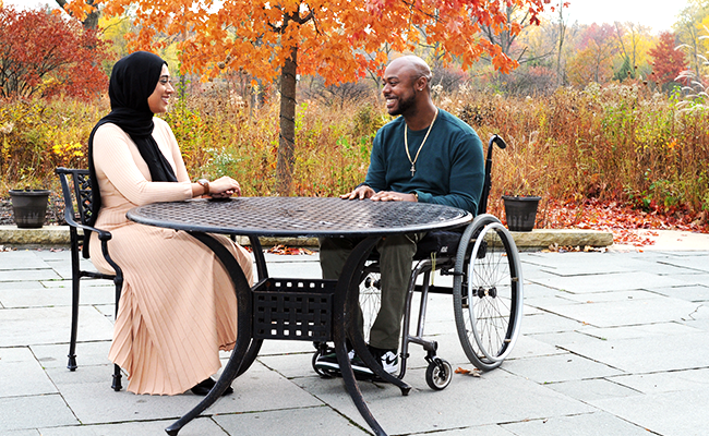 A woman and man are sitting at a table outdoors. The woman is wearing a black headscarf and a pink dress and the man is wearing a blue jacket and is sitting in a wheelchair.