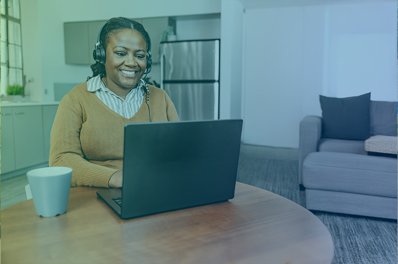A woman wearing an brown V-neck sweater is seated at a table and typing on a laptop. She is smiling and wearing a headset.